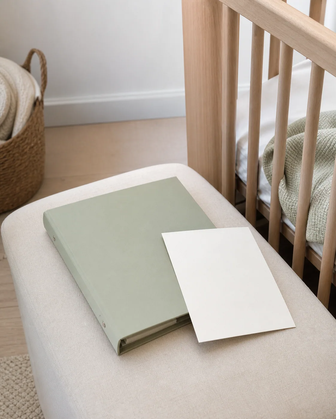 A slim sage binder and blank letter card beside a contemporary crib in natural daylight.
