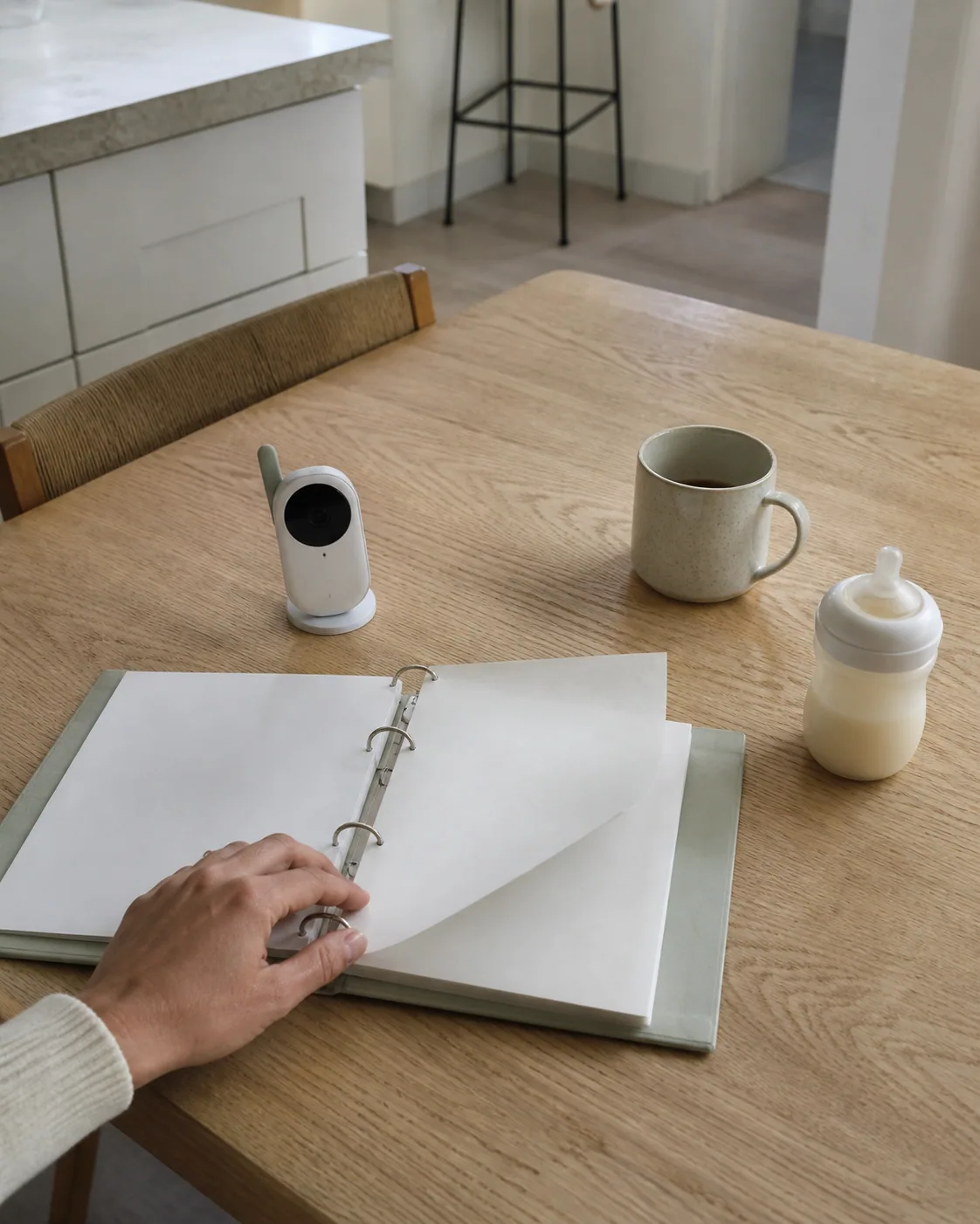 A hand turning a blank binder page beside a baby monitor and mug at a current kitchen table.